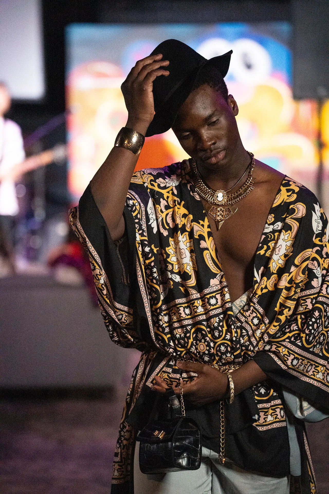 A model in an open black and gold baroque-print shirt, tipping his hat.