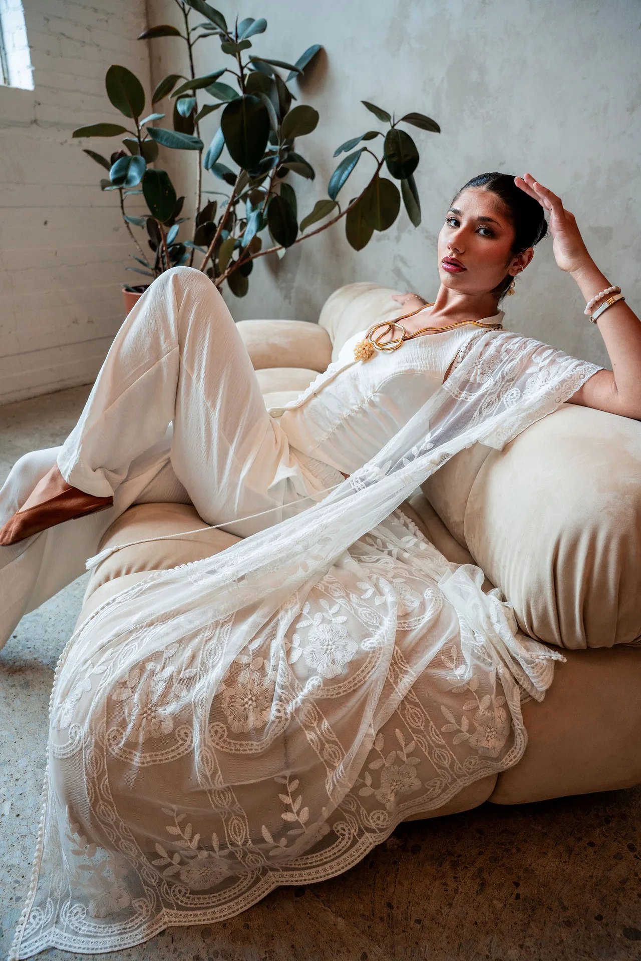 A model in a white lace duster and trousers reclining on a cream sofa beneath a rubber plant.