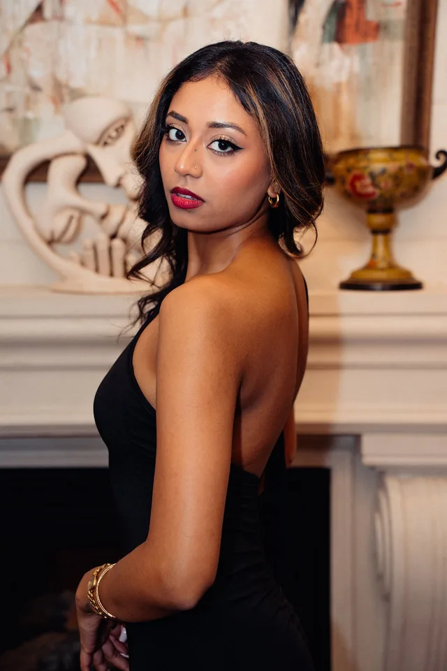 Portrait of a guest in a black halter dress with a red lip beside a marble fireplace.