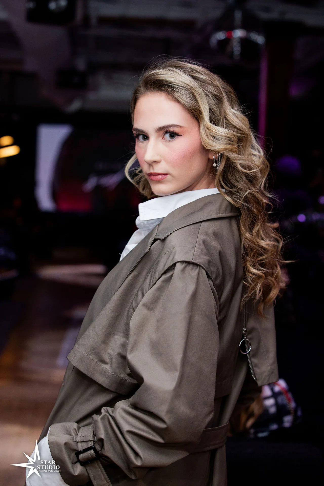 Portrait of a model in an olive trench coat over a white shirt.