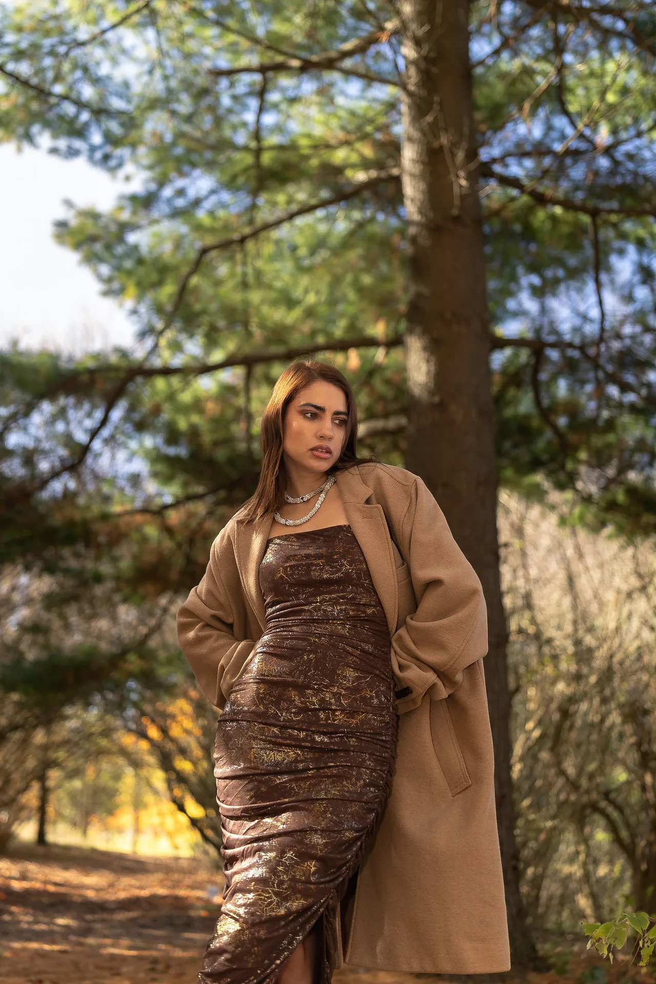 A model in a bronze ruched dress and camel coat among tall pines.