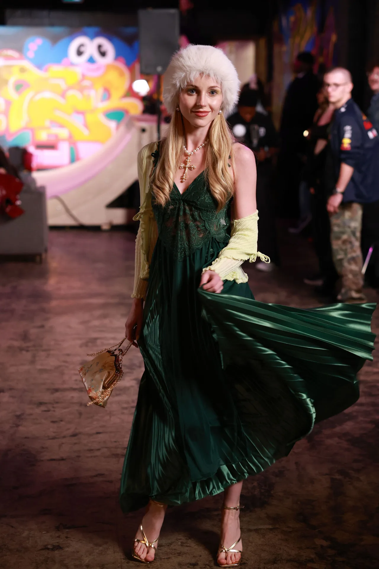 A model in an emerald pleated dress and white fur hat, skirt swinging, in front of a graffiti set.
