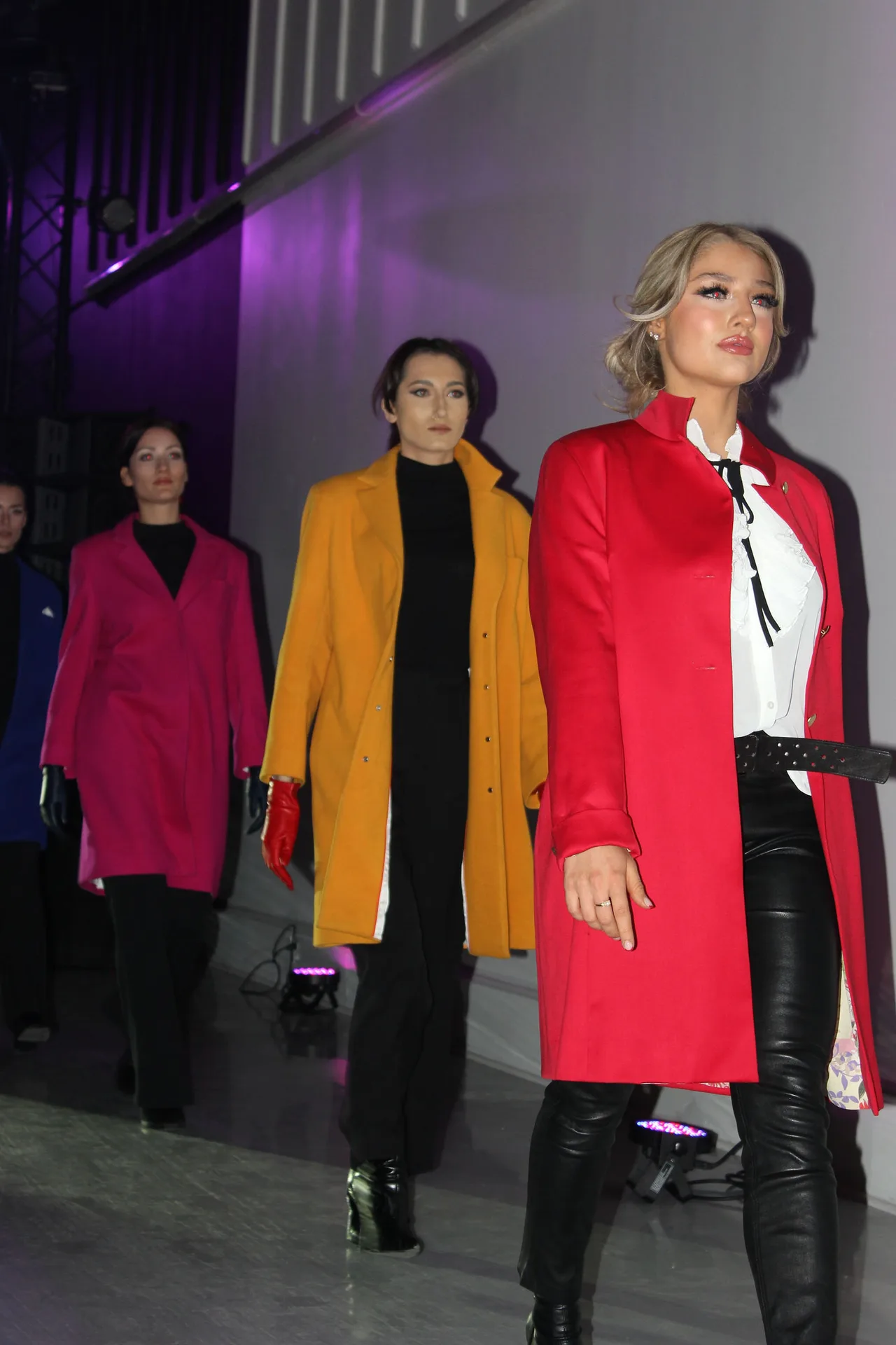 Three models in pink, mustard and red coats walking the runway together.