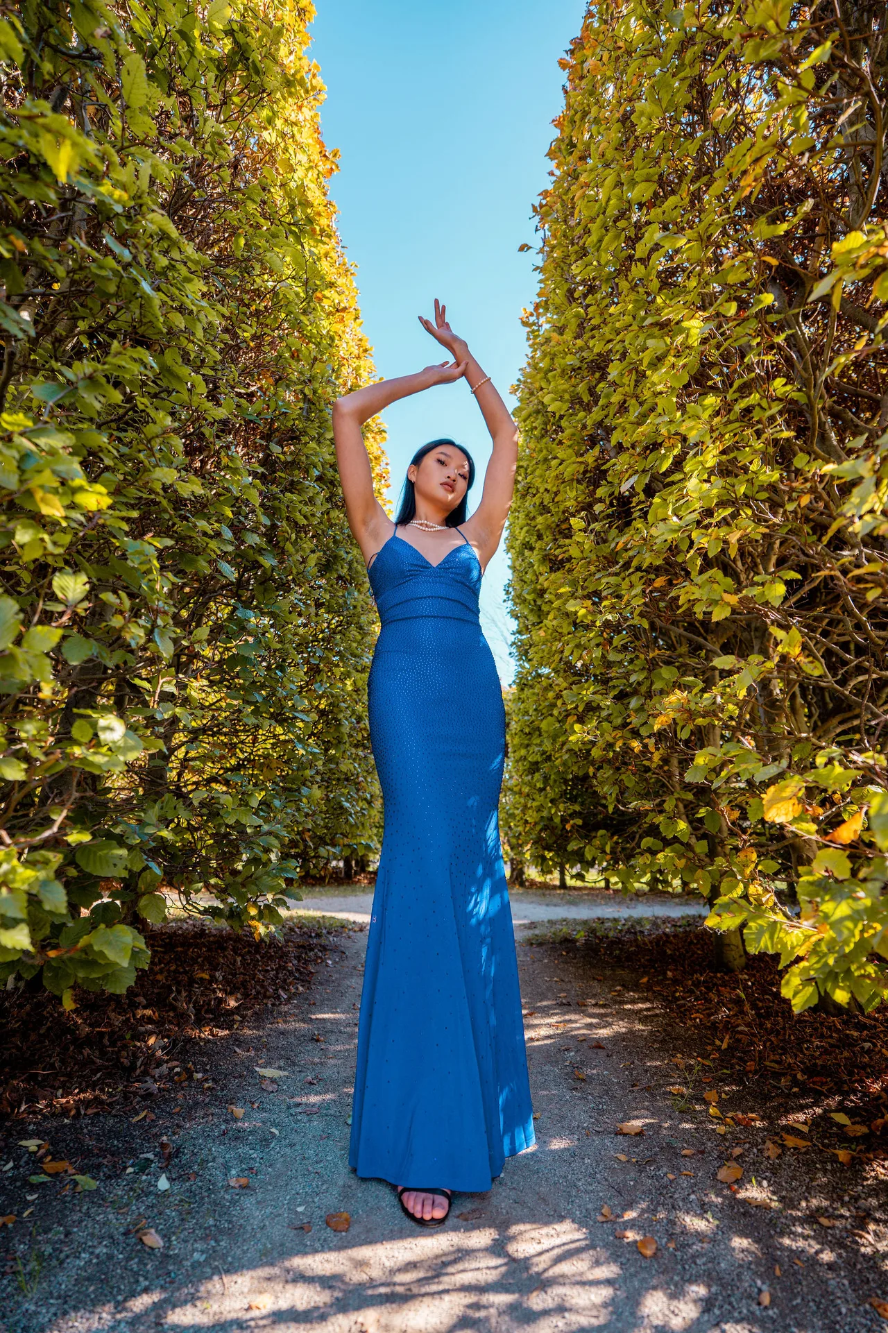 A model in a royal blue satin gown, arms raised, between two tall hedges.