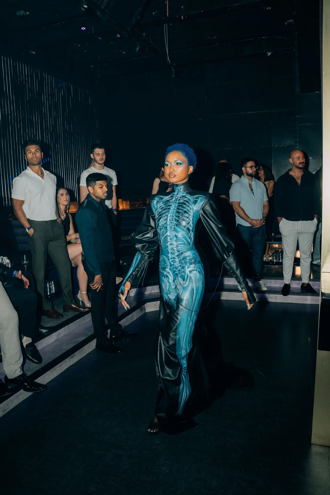 A model in a skeleton-print catsuit with blue hair walking between guests.