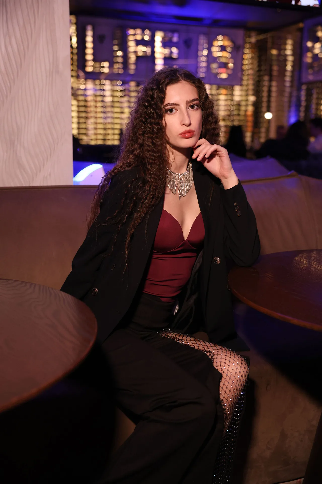 A guest in a burgundy top, black blazer and pearls against a neon-lit wall.