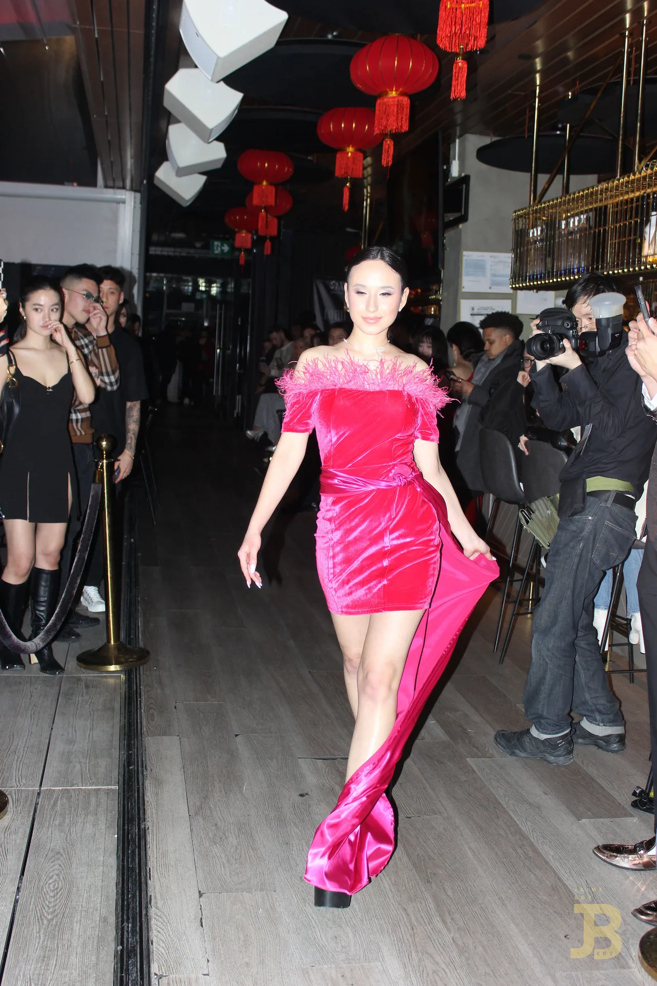 A model in a hot pink velvet dress with a long train walking the runway under red lanterns.