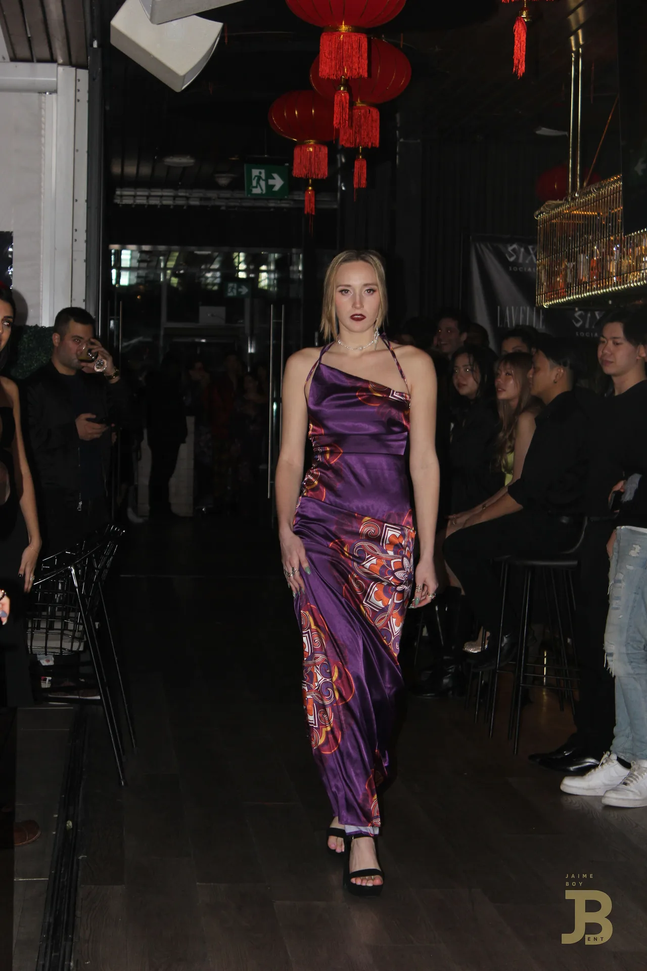 A model in a purple printed one-shoulder gown walking between guests.