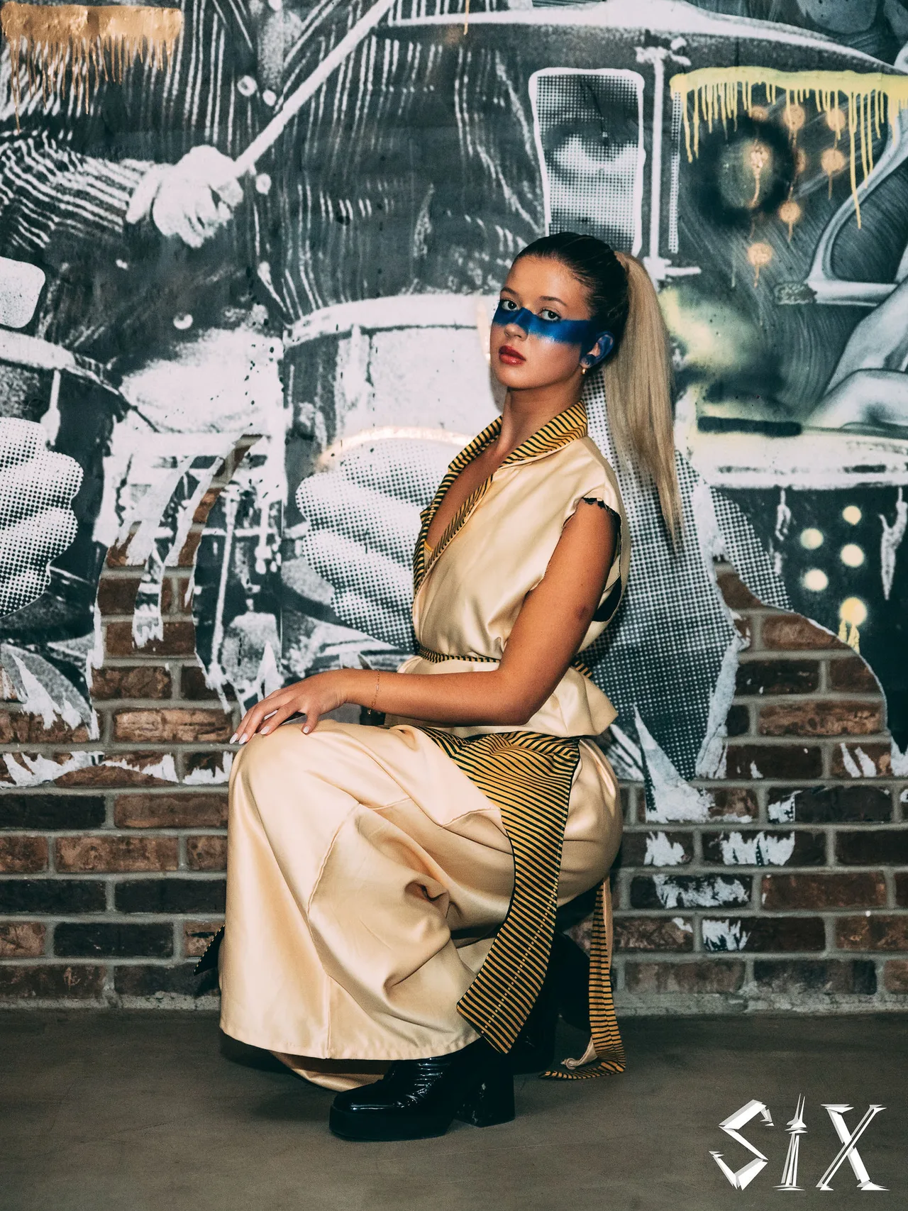 A model in a cream suit crouching in front of a graffiti mural, with blue eye paint.