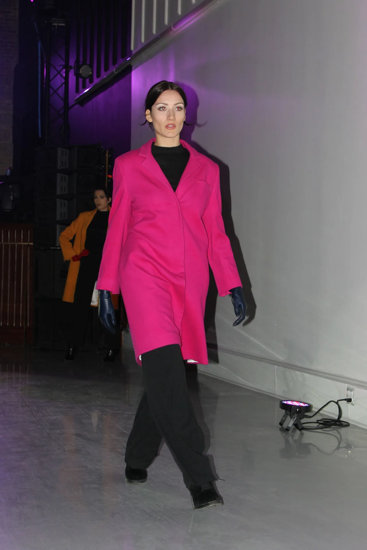 A model in a hot pink coat and black trousers walking a purple-lit runway.