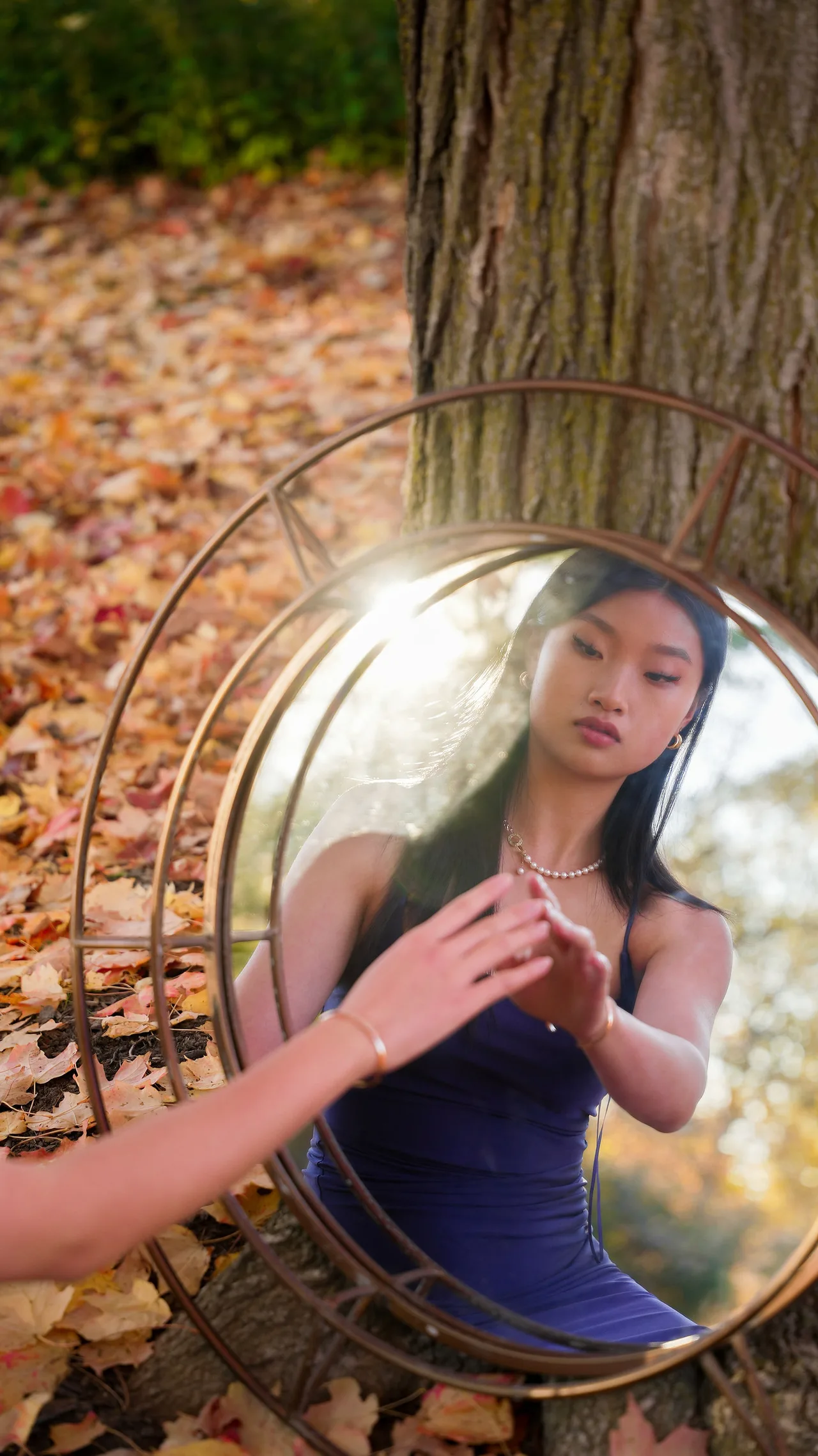 A model in a purple dress reflected in a round mirror propped against a tree.