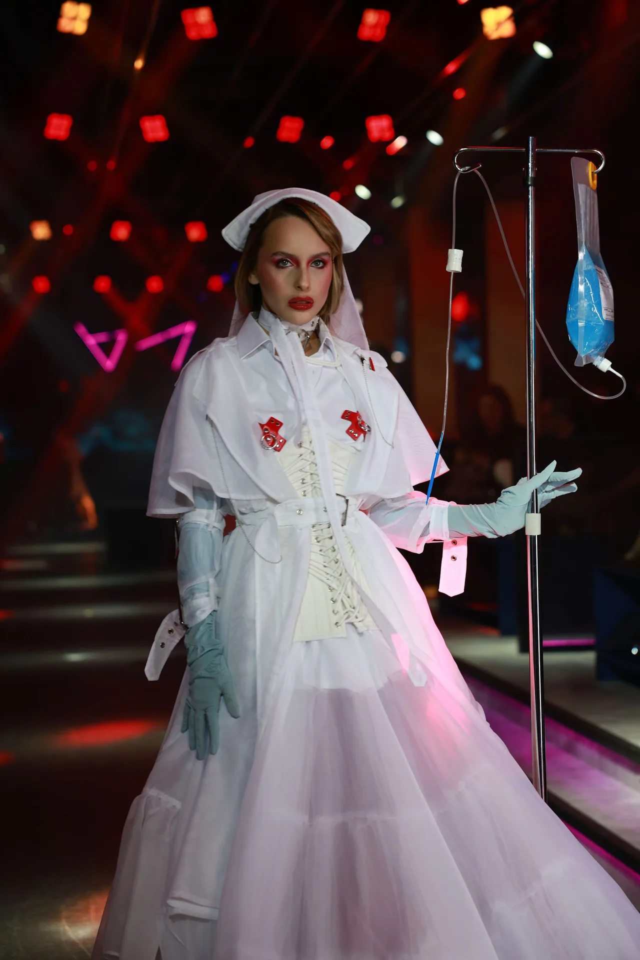 A model in a white Victorian-style gown and nurse's cap beside an IV stand, under red runway lights.