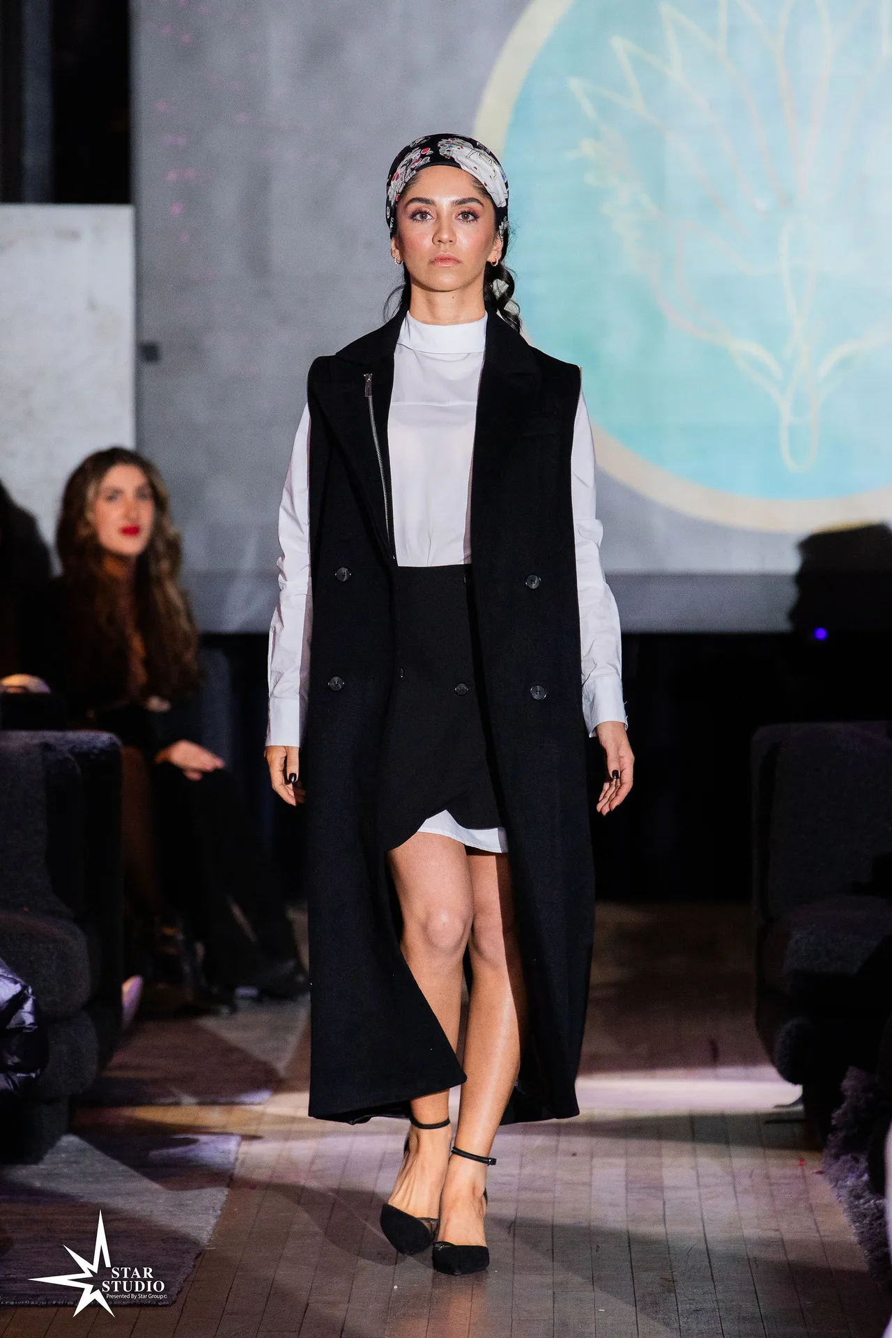 A model in a long black vest coat over a white shirt, with a printed headscarf.