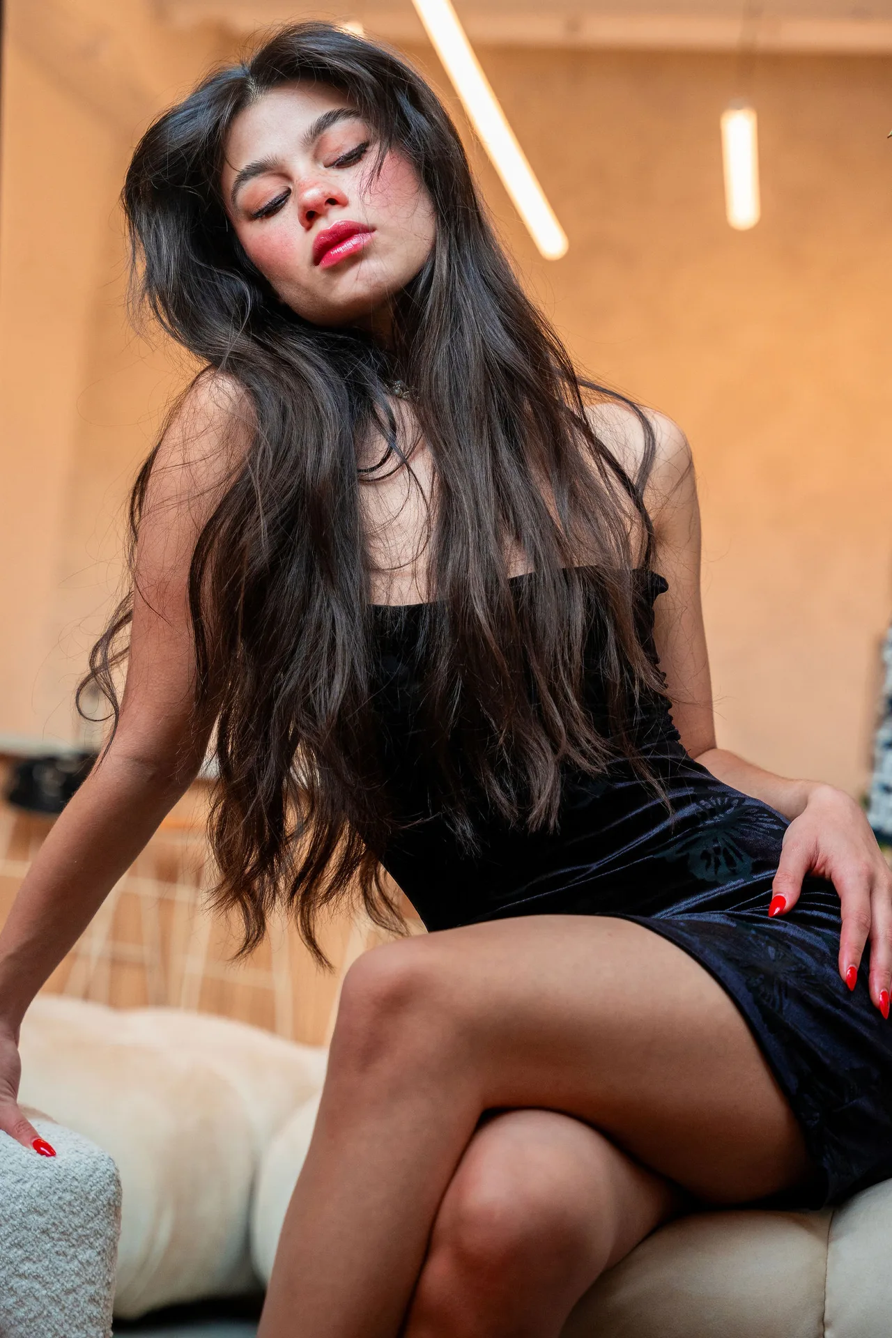 A model in a black mini dress seated in warm light, eyes closed, long hair past her shoulders.
