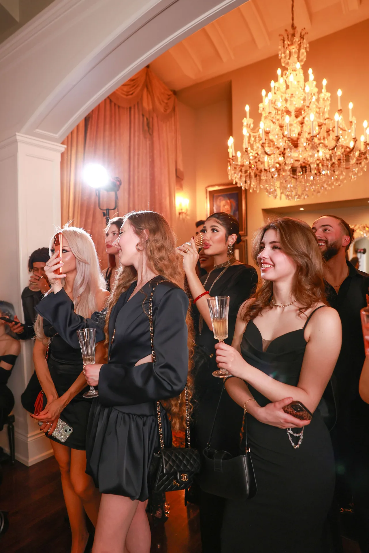 Guests in black evening wear gathered beneath a chandelier in the mansion.