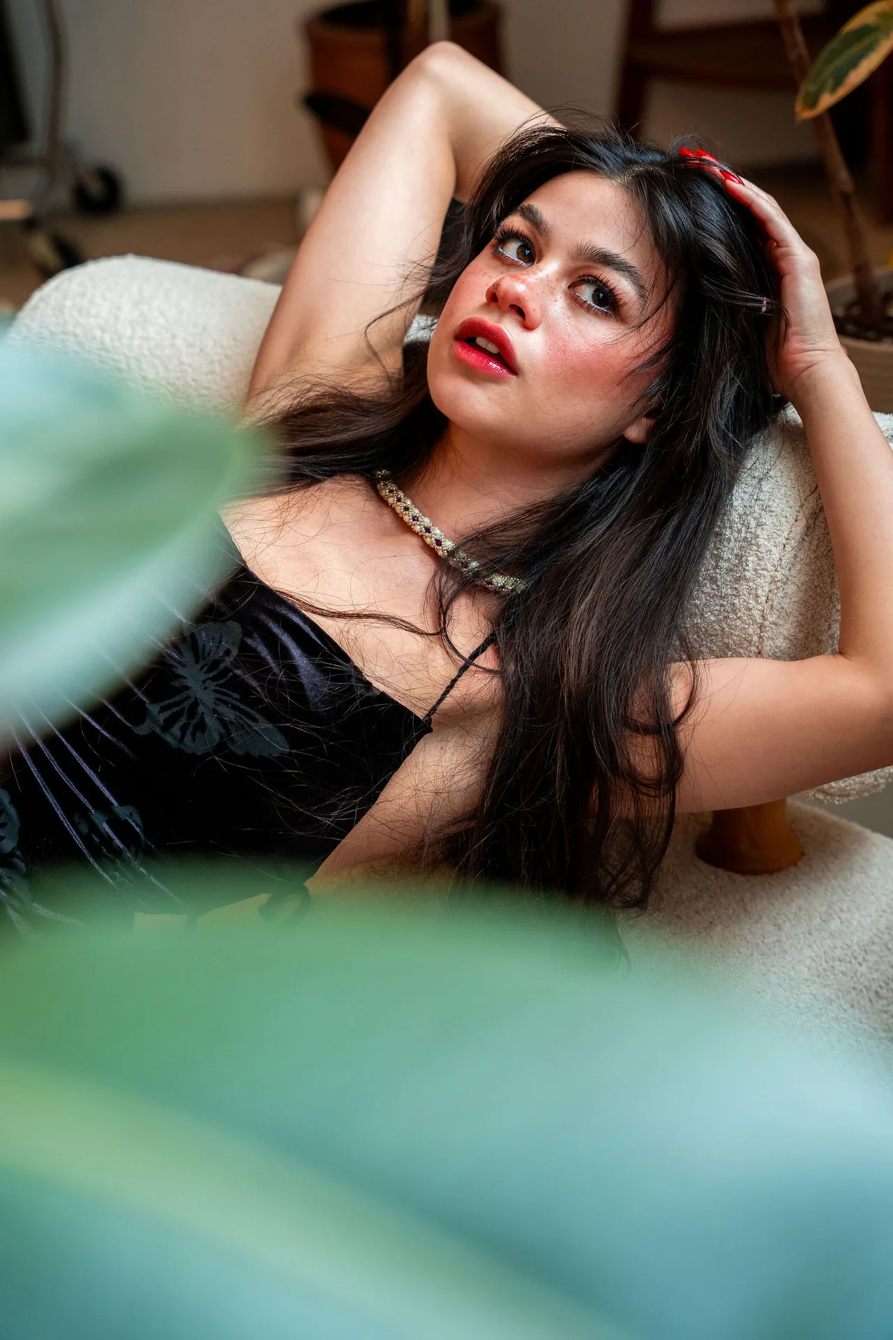 A model in a black velvet dress reclining on a boucle chair, framed by out-of-focus leaves.