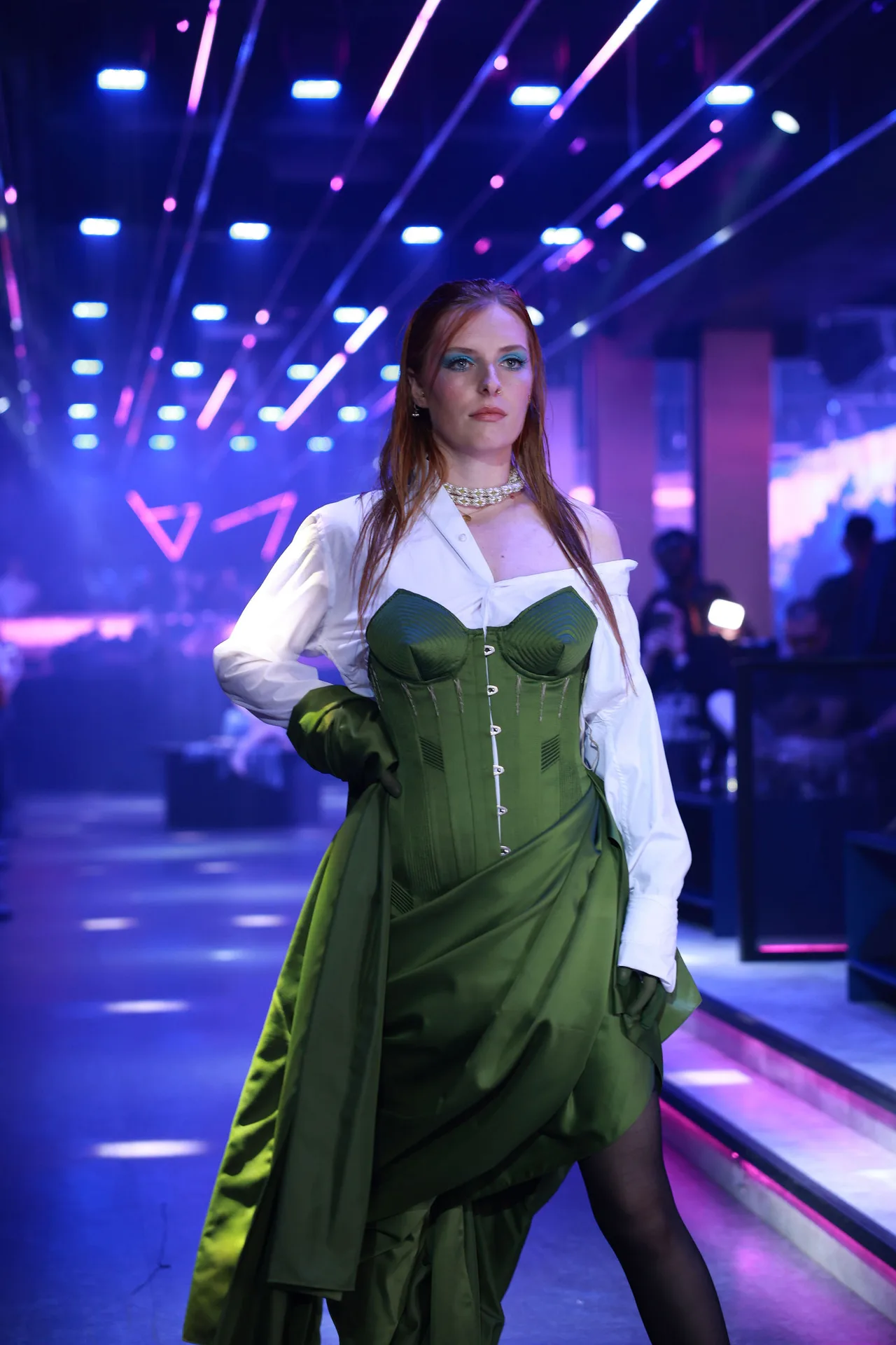 A model in an emerald corset gown over a white shirt on a blue-lit runway.