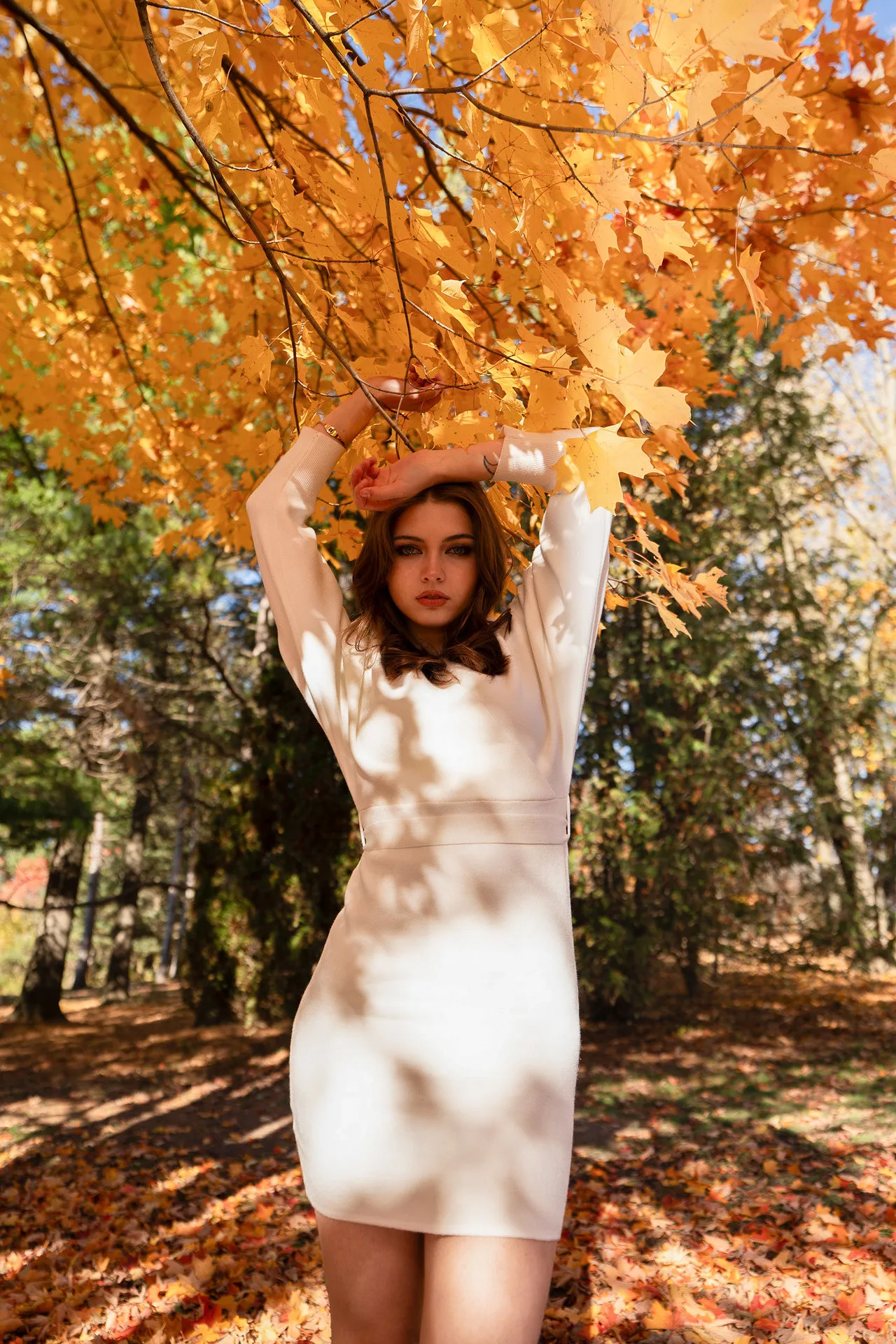 A model in a cream sweater dress with arms raised under orange maple leaves.