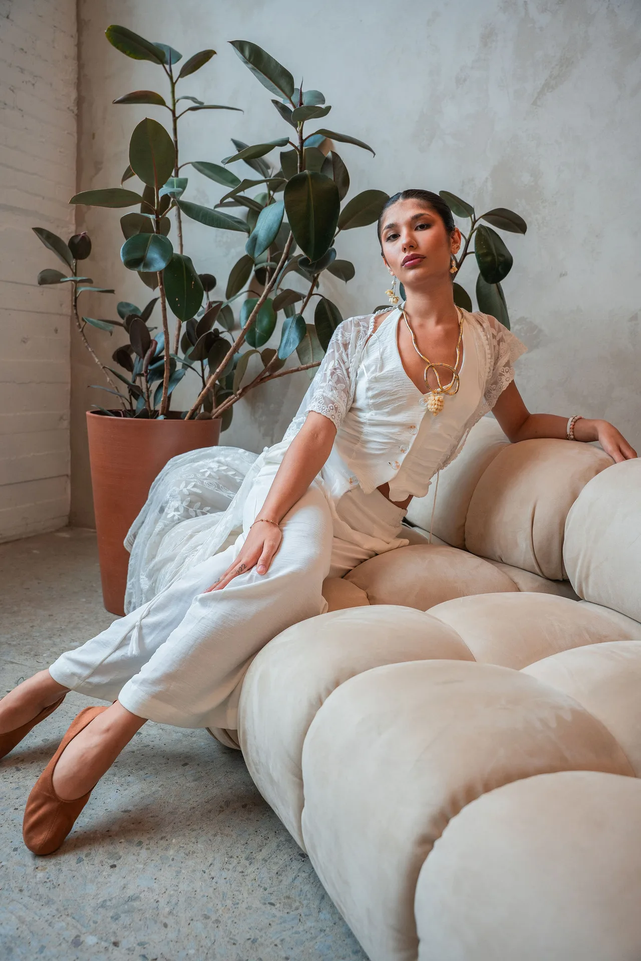 A model in white lace lounging across a cream modular sofa in a bright studio with plants.