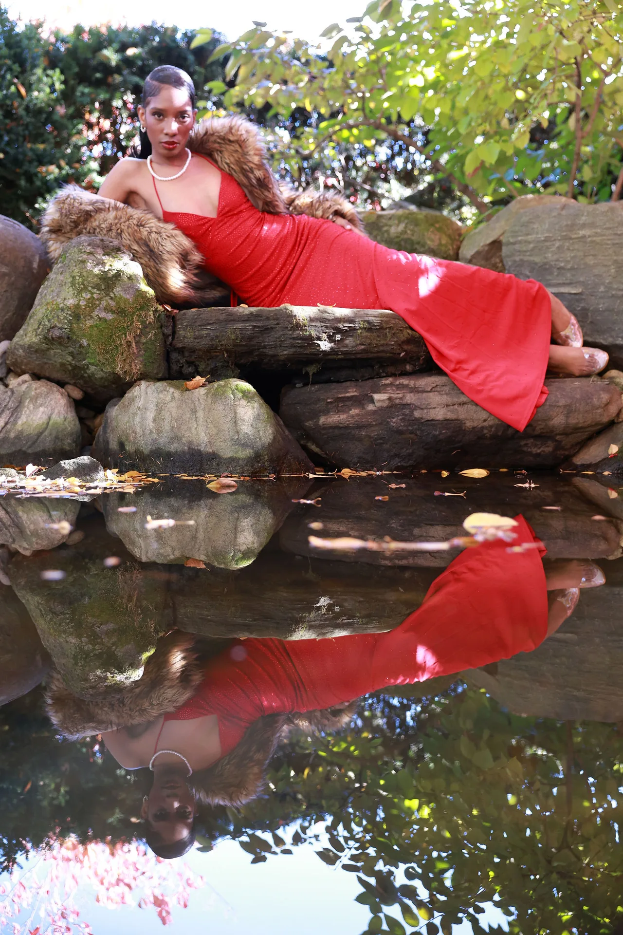 A model in a red gown reclining on rocks, mirrored in the water below.