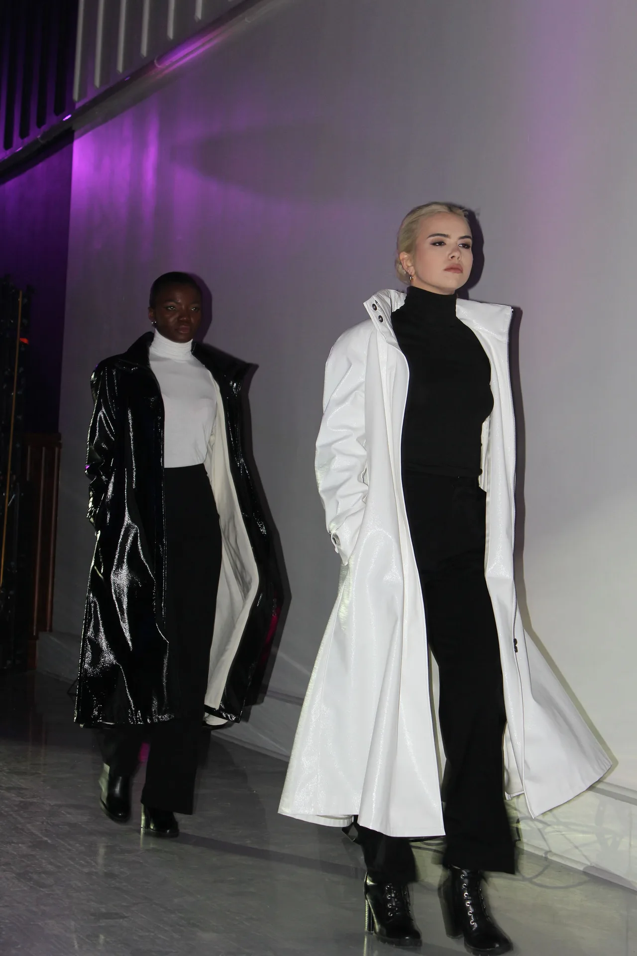 Two models in long black patent and white coats on the runway.